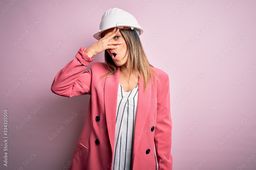 Wall mural Young beautiful brunette architect woman wearing safety helmet over pink background peeking in shock covering face and eyes with hand, looking through fingers with embarrassed expression.