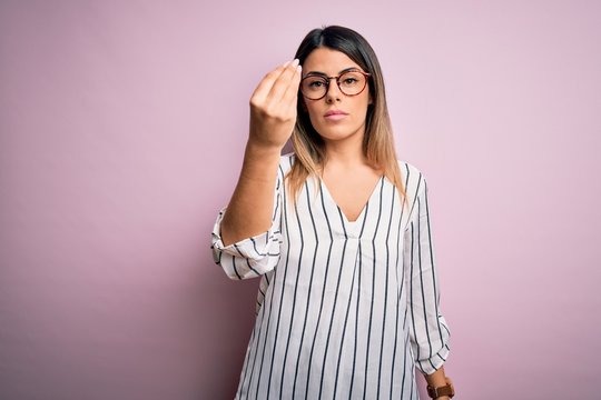 Young beautiful woman wearing casual striped t-shirt and glasses over pink background Doing Italian gesture with hand and fingers confident expression