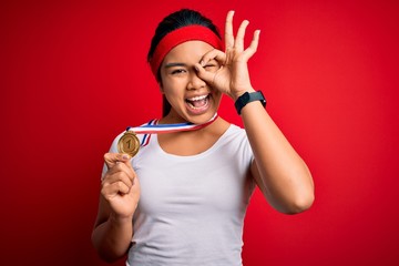 Young asian champion girl winning medal standing over isolated red background with happy face smiling doing ok sign with hand on eye looking through fingers