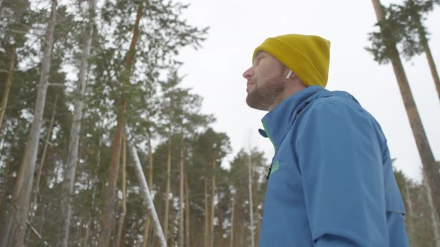 Slowmo tracking shot of handsome man in blue windbreaker and yellow hat standing in forest in cold winter morning and looking around, then starting running