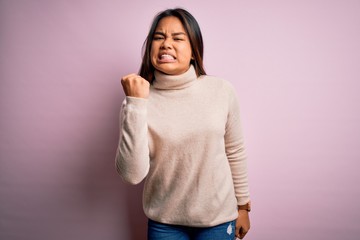 Young beautiful asian girl wearing casual turtleneck sweater over isolated pink background angry and mad raising fist frustrated and furious while shouting with anger. Rage and aggressive concept.