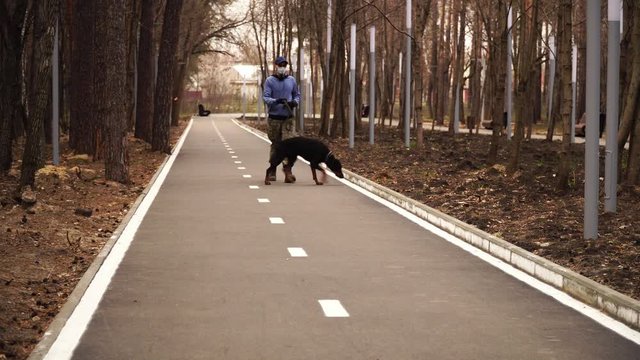 Man in medical mask is walking a big black dog in desert park Theme of the viral disease pandemic and urban quarantine. The owner in camouflage pants walks a pet in a uninhabited city.