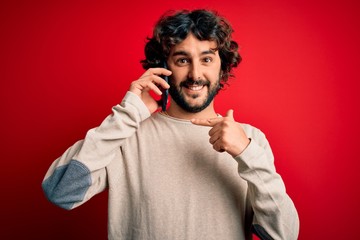 Handsome man with beard having conversation talking on the smartphone over red background very happy pointing with hand and finger
