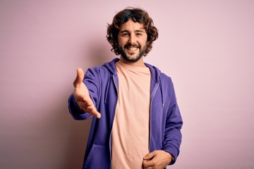 Young handsome sporty man with beard wearing casual sweatshirt over pink background smiling cheerful offering palm hand giving assistance and acceptance.