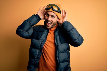 Young handsome skier man with beard wearing snow sportswear and ski goggles Smiling cheerful playing peek a boo with hands showing face. Surprised and exited