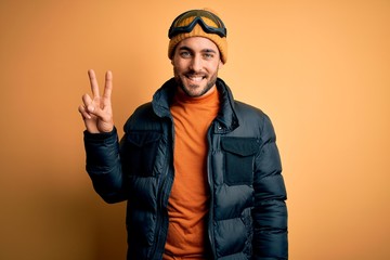 Young handsome skier man with beard wearing snow sportswear and ski goggles smiling looking to the camera showing fingers doing victory sign. Number two.