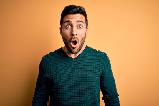Young handsome man with beard wearing casual sweater standing over yellow background afraid and shocked with surprise expression, fear and excited face.