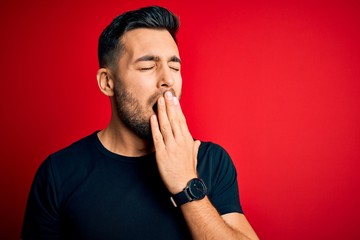 Young handsome man wearing casual black t-shirt standing over isolated red background bored yawning...