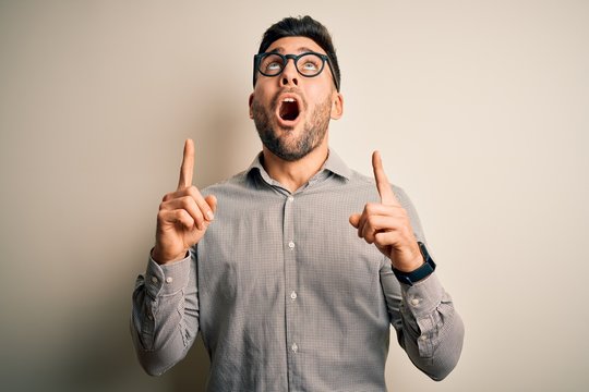 Young handsome man wearing elegant shirt and glasses over isolated white background amazed and surprised looking up and pointing with fingers and raised arms.