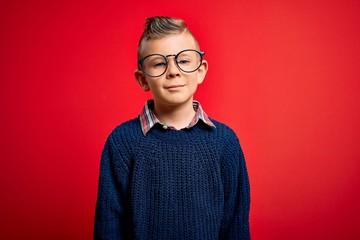 Young little caucasian kid with blue eyes standing wearing smart glasses over red background with serious expression on face. Simple and natural looking at the camera.