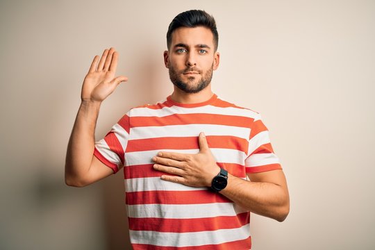 Young handsome man wearing casual striped t-shirt standing over isolated white background Swearing with hand on chest and open palm, making a loyalty promise oath