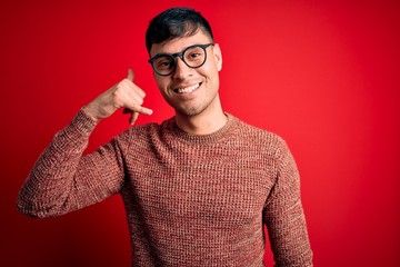 Young handsome hispanic man wearing nerd glasses over red background smiling doing phone gesture with hand and fingers like talking on the telephone. Communicating concepts.