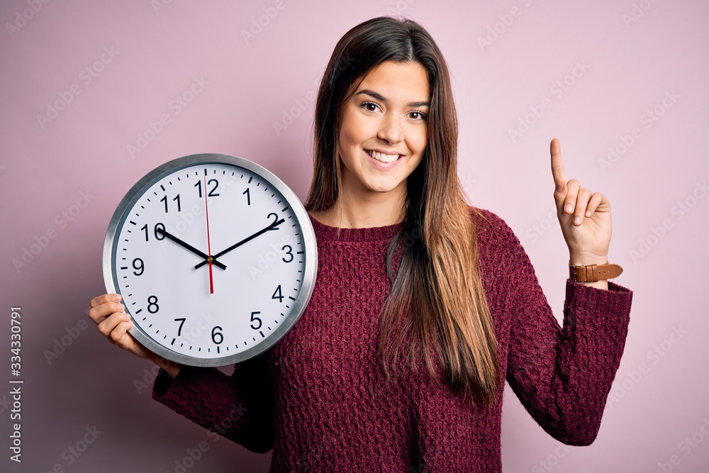 Sticker Young beautiful girl doing countdown holding big clock over isolated pink background surprised with an idea or question pointing finger with happy face, number one