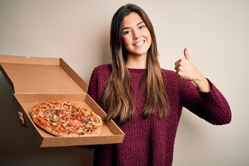 Young beautiful girl holding delivery box with Italian pizza standing over white background happy with big smile doing ok sign, thumb up with fingers, excellent sign