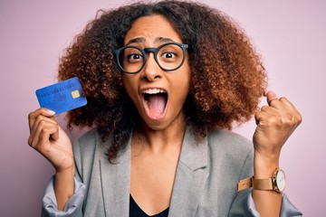 Young african american businesswoman with afro hair holding credit card over pink background...