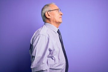 Grey haired senior business man wearing glasses standing over purple isolated background looking to side, relax profile pose with natural face with confident smile.