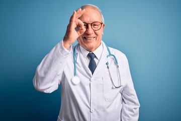 Senior grey haired doctor man wearing stethoscope and medical coat over blue background doing ok gesture with hand smiling, eye looking through fingers with happy face.