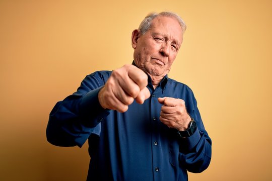 Grey haired senior man wearing casual blue shirt standing over yellow background Punching fist to fight, aggressive and angry attack, threat and violence