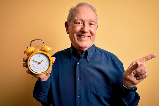 Senior grey haired man holding vintage alarm clock over yellow background very happy pointing with hand and finger to the side