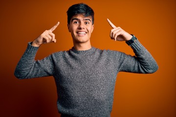 Young handsome man wearing casual sweater standing over isolated orange background smiling pointing to head with both hands finger, great idea or thought, good memory