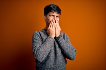 Young handsome man wearing casual sweater standing over isolated orange background laughing and embarrassed giggle covering mouth with hands, gossip and scandal concept