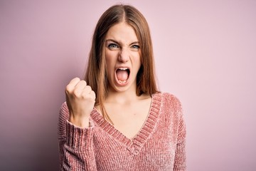 Young beautiful redhead woman wearing casual sweater over isolated pink background angry and mad raising fist frustrated and furious while shouting with anger. Rage and aggressive concept.