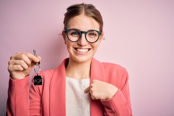 Young beautiful redhead house agent woman holding home key over pink bakcground with surprise face pointing finger to himself