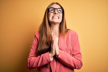 Young beautiful redhead woman wearing jacket and glasses over isolated yellow background begging...