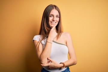 Obraz premium Young beautiful redhead woman wearing casual t-shirt over isolated yellow background looking confident at the camera smiling with crossed arms and hand raised on chin. Thinking positive.