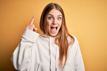 Young beautiful redhead sporty woman wearing sweatshirt over isolated yellow background pointing finger up with successful idea. Exited and happy. Number one.