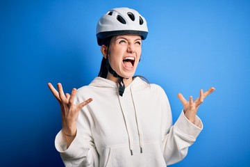 Young beautiful redhead cyclist woman wearing bike helmet over isolated blue background crazy and mad shouting and yelling with aggressive expression and arms raised. Frustration concept.