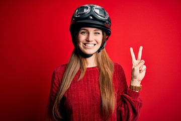 Young beautiful redhead motocyclist woman wearing moto helmet over red background smiling with happy face winking at the camera doing victory sign. Number two.