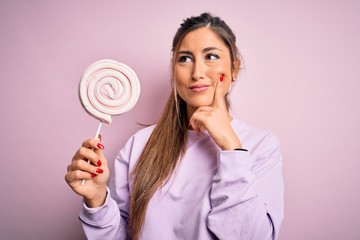 Young beautiful brunette woman eating sweet candy over isolated pink background serious face...
