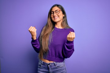 Young beautiful smart woman wearing glasses over purple isolated background very happy and excited doing winner gesture with arms raised, smiling and screaming for success. Celebration concept.