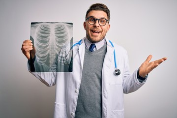 Young doctor man wearing medical coat looking at chest scan radiography over isolated background very happy and excited, winner expression celebrating victory screaming with big smile and raised hands