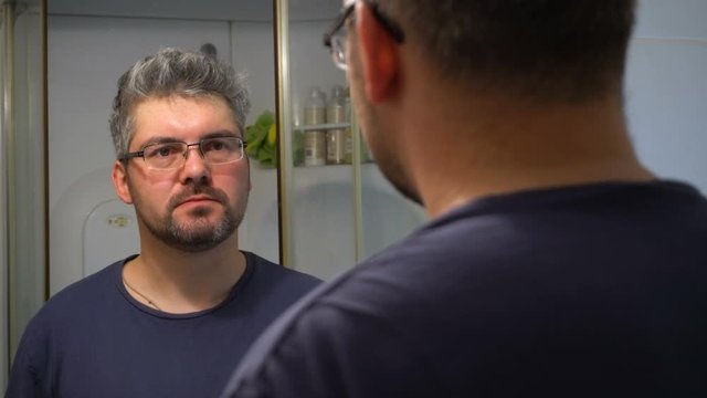 A man in glasses with a beard in the bathroom. It twists its face. Changes emotions. Reflection in the mirror. Morning Awakening.