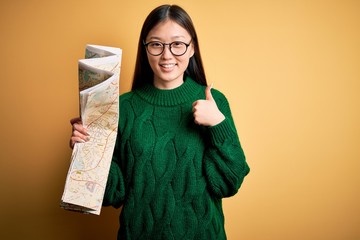 Young asian turist woman looking at city tourist map on a trip over yellow background happy with big smile doing ok sign, thumb up with fingers, excellent sign