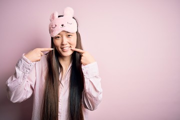 Young asian woman wearing pajama and sleep mask over pink isolated background smiling cheerful showing and pointing with fingers teeth and mouth. Dental health concept.