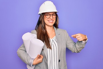Young hispanic architect woman wearing builder hat and holding blueprints over yellow background with surprise face pointing finger to himself