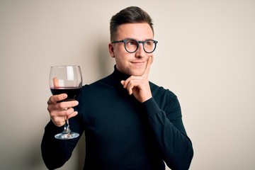 Young handsome caucasian man drinking an alcoholic glass of red wine over isolated background serious face thinking about question, very confused idea