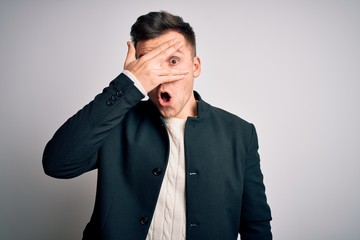 Young handsome business mas wearing elegant winter coat standing over isolated background peeking in shock covering face and eyes with hand, looking through fingers with embarrassed expression.
