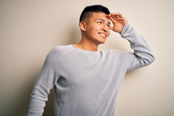 Young handsome latin man wearing casual sweater standing over isolated white background very happy and smiling looking far away with hand over head. Searching concept.