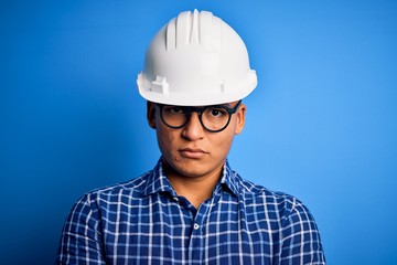 Young handsome engineer latin man wearing safety helmet over isolated blue background with serious expression on face. Simple and natural looking at the camera.