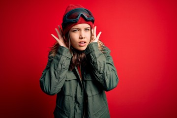 Young blonde girl wearing ski glasses and winter coat for ski weather over red background Trying to hear both hands on ear gesture, curious for gossip. Hearing problem, deaf