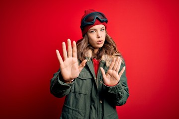Young blonde girl wearing ski glasses and winter coat for ski weather over red background Moving away hands palms showing refusal and denial with afraid and disgusting expression. Stop and forbidden.