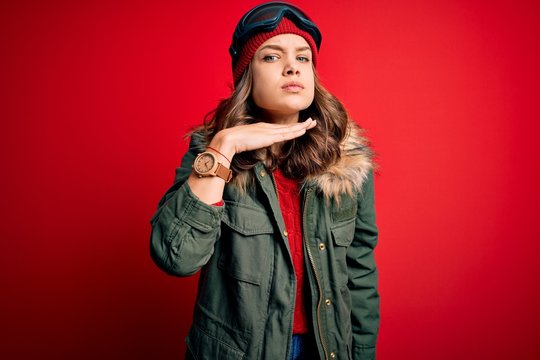 Young blonde girl wearing ski glasses and winter coat for ski weather over red background cutting throat with hand as knife, threaten aggression with furious violence