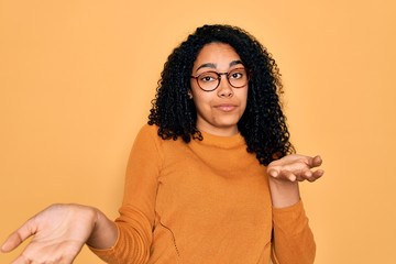 Young african american woman wearing casual sweater and glasses over yellow background clueless and...