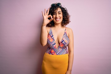 Young beautiful arab woman on vacation wearing swimsuit and sunglasses over pink background smiling positive doing ok sign with hand and fingers. Successful expression.