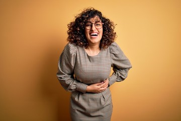 Beautiful arab business woman wearing dress and glasses standing over yellow background smiling and laughing hard out loud because funny crazy joke with hands on body.
