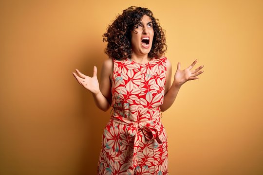 Young beautiful curly arab woman on vacation wearing summer floral dress and sunglasses crazy and mad shouting and yelling with aggressive expression and arms raised. Frustration concept.